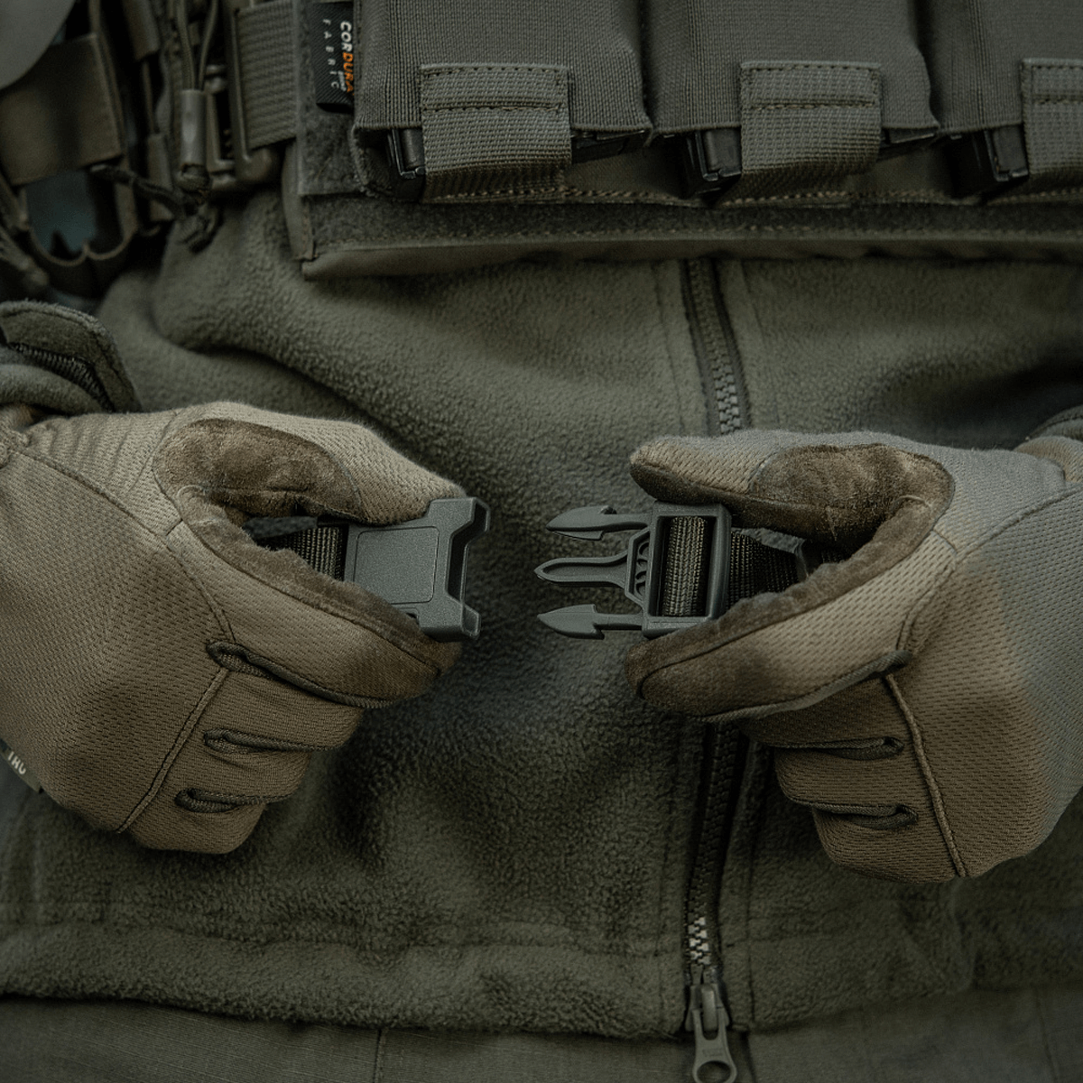 M-Tac Siddeunderlag med bælte i brug af person iført militærjakke og handsker, der viser spænde detaljer.