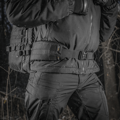 M-Tac Rygsæk Gen.II Elite Large båret af en person i vintertøj i en skov, fremstillet af slidstærk Cordura for maksimal holdbarhed.