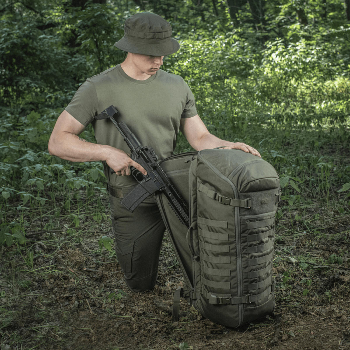 M-Tac Rygsæk Gen.II Elite Large i skoven med våben, viser holdbar konstruktion og funktionalitet til udendørs eventyr.