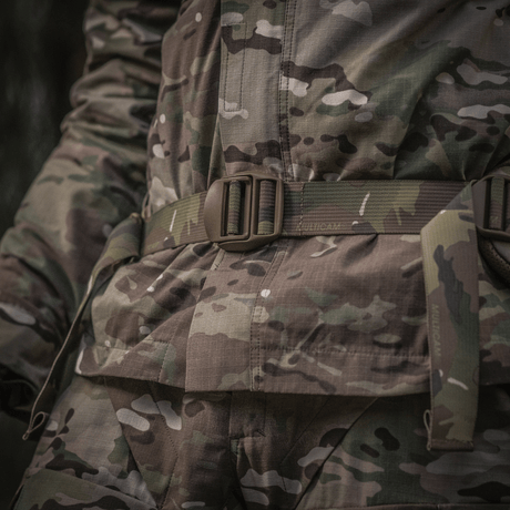 Nærbillede af militær uniform med camouflagemønster, fokuseret på bælteområdet i skovmiljø.