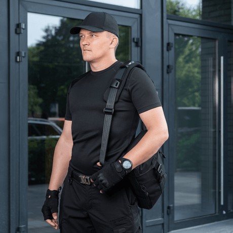 Mand bærer taske med M‑Tac Skulderremspude 50 mm Elite for øget komfort og støtte, iført sort tøj foran moderne bygning.