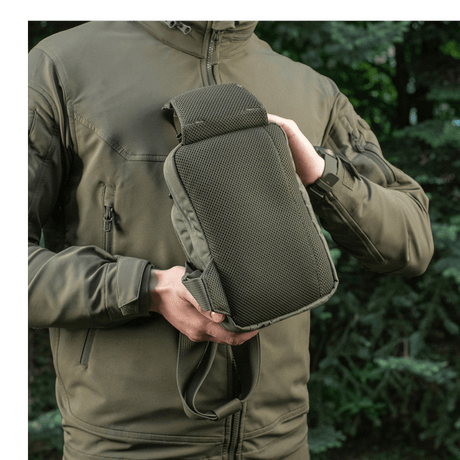 Person viser M-Tac Sling Pistoltaske Elite Hex udendørs i skovmiljø, iført jakke med lynlås.