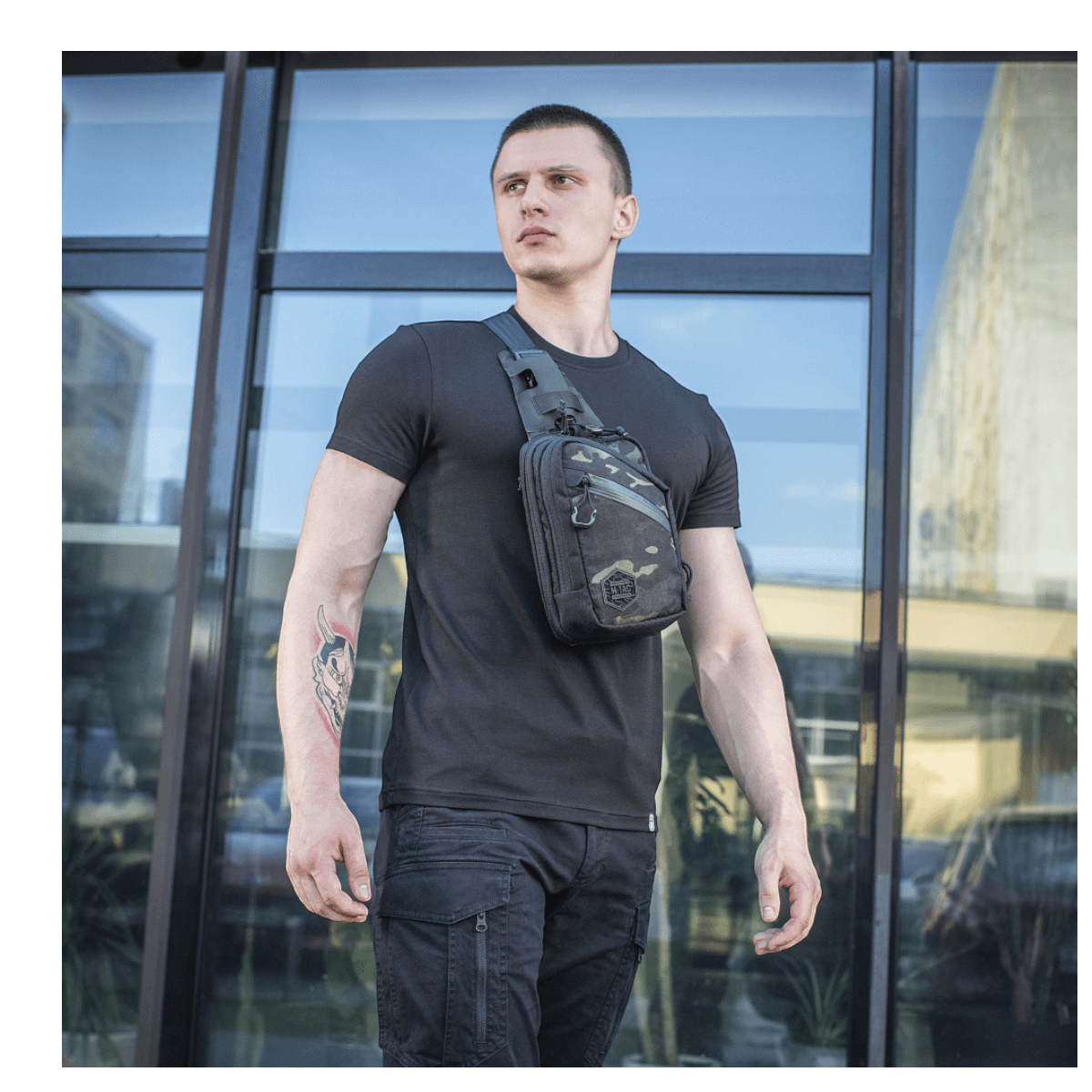 Mand iført M-Tac Sling Pistoltaske Elite Hex foran moderne bygning i sort tøj, fremviser taske med justerbar rem og rummelige rum.