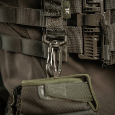 M-Tac Karabinhage Tilbehør fastgjort til MOLLE-system på taktisk vest, med justerbar Velcro-rem for ekstra udstyrssikring.