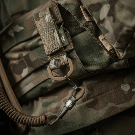 M-Tac Fastgørelse med D-ring monteret på camo-rygsæk, ideel til taktisk brug med MOLLE-system. Velcro-lukning, kompakt og letvægtsdesign.