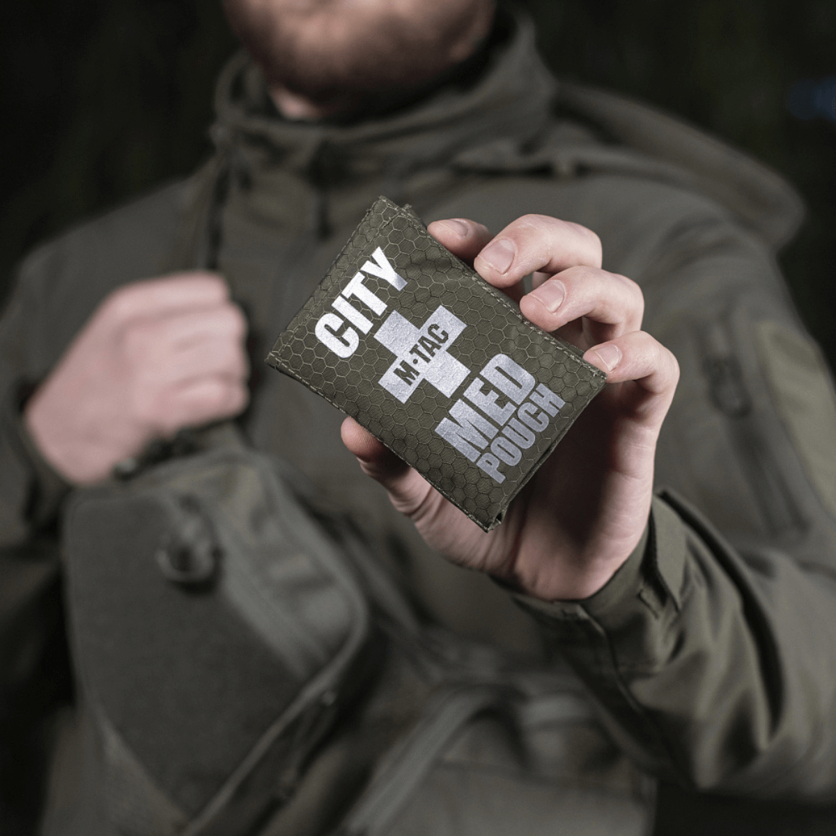 Mand holder M-Tac City Med Pouch Hex, kompakt førstehjælpskit i slidstærkt Hex Polyester med medicinsk kors og Velcro-lukning.