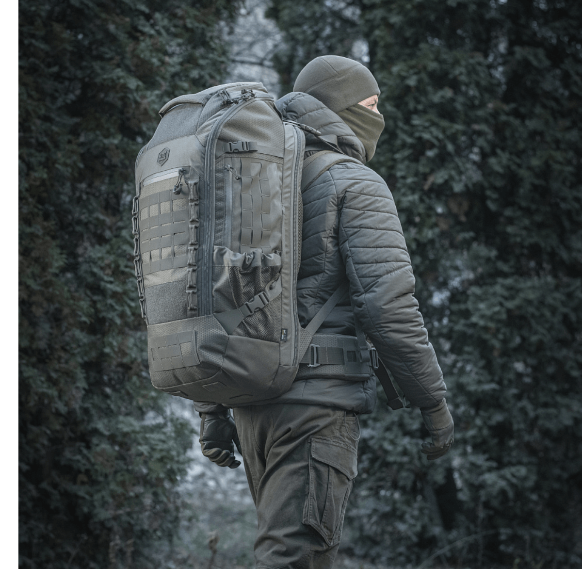 M-Tac Rygsæk Large Elite Hex Gen. IV i et skovmiljø, designet til bæring af våben og beskyttelse med holdbart Cordura-materiale.