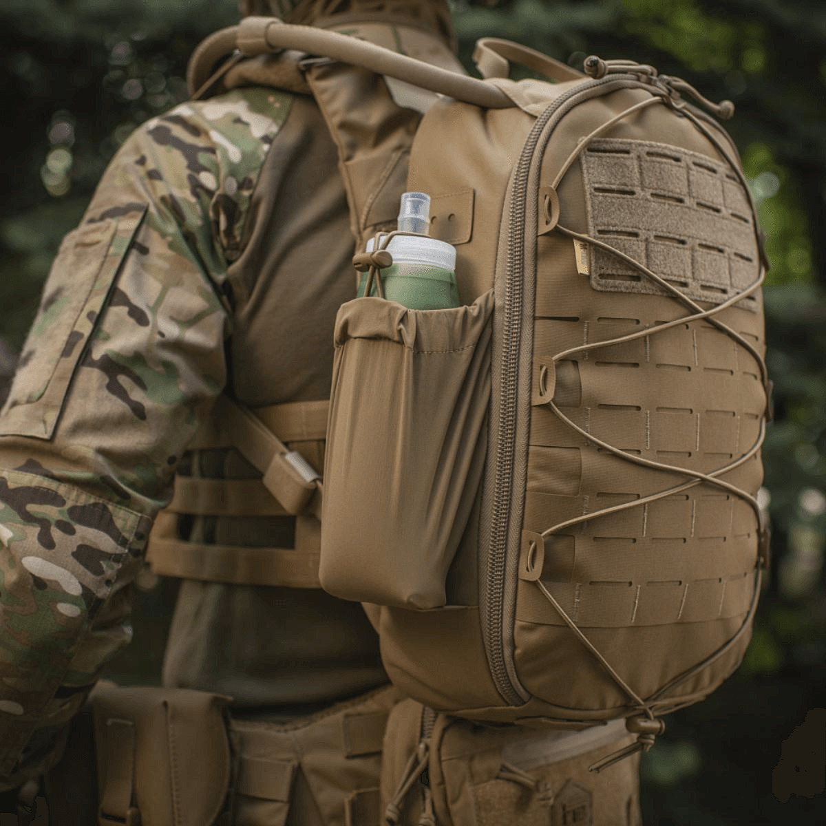 Taktisk militærrygsæk med vandflaske i sidelommen, set udendørs. Velegnet til udendørs aktiviteter og kampagner.