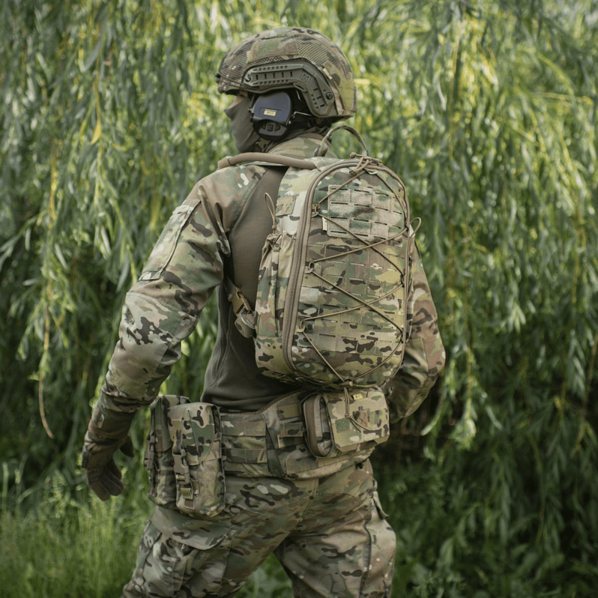 Soldat i camouflageuniform med rygsæk, set bagfra i en skov.