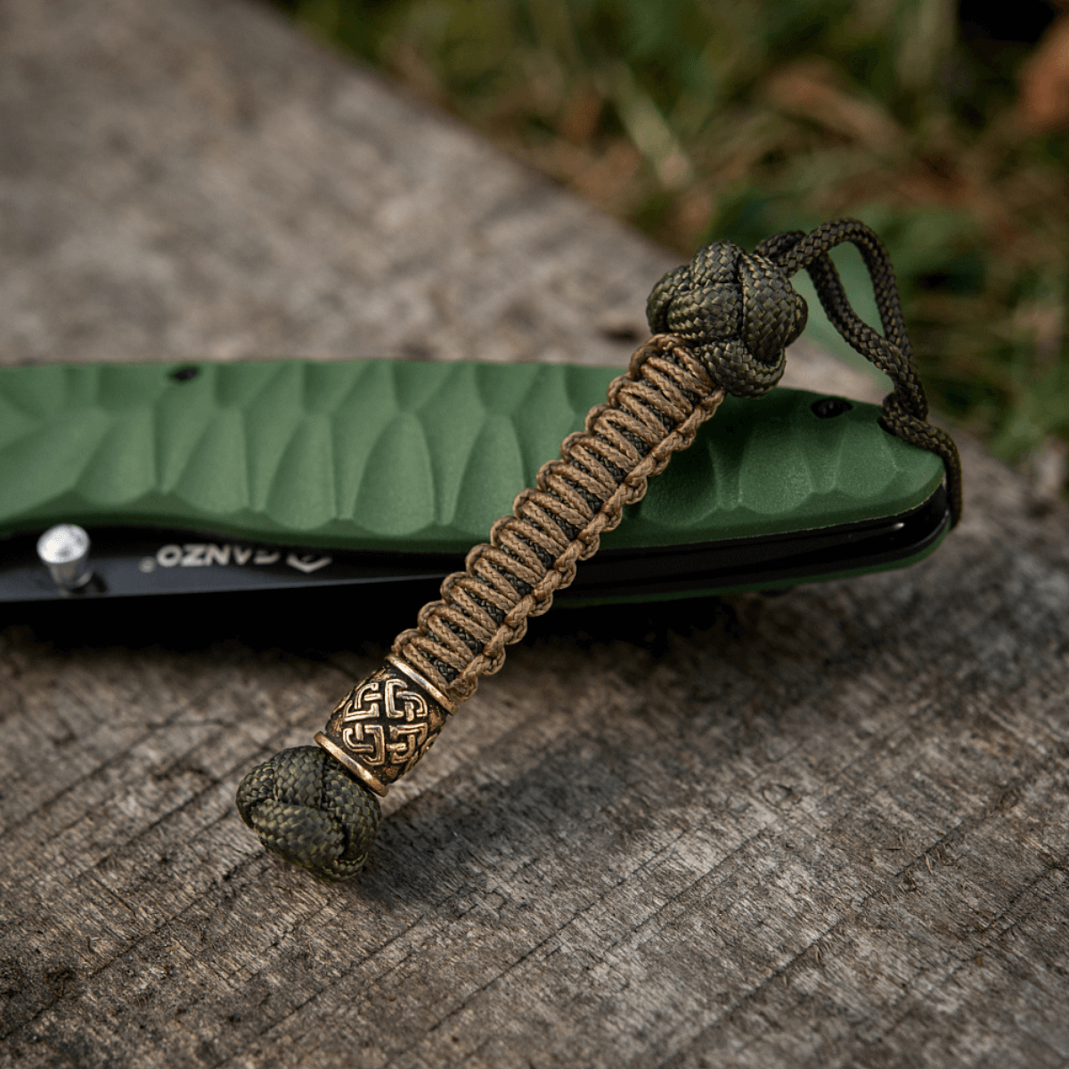 M-Tac Knife Lanyard Celtic i flettet paracord ved siden af en grøn lommekniv på træoverflade
