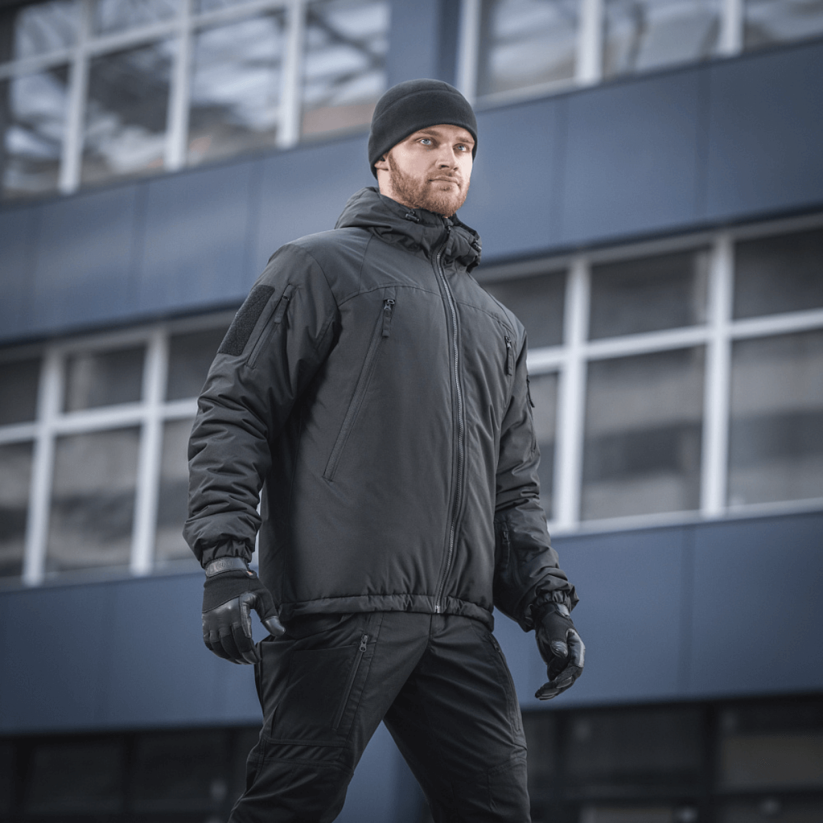Mand iført M-Tac Alpha Gen.III Pro Vinterjakke med justerbar hætte, perfekt til koldt vejr og hverdagsbrug, baggrund med moderne bygning.