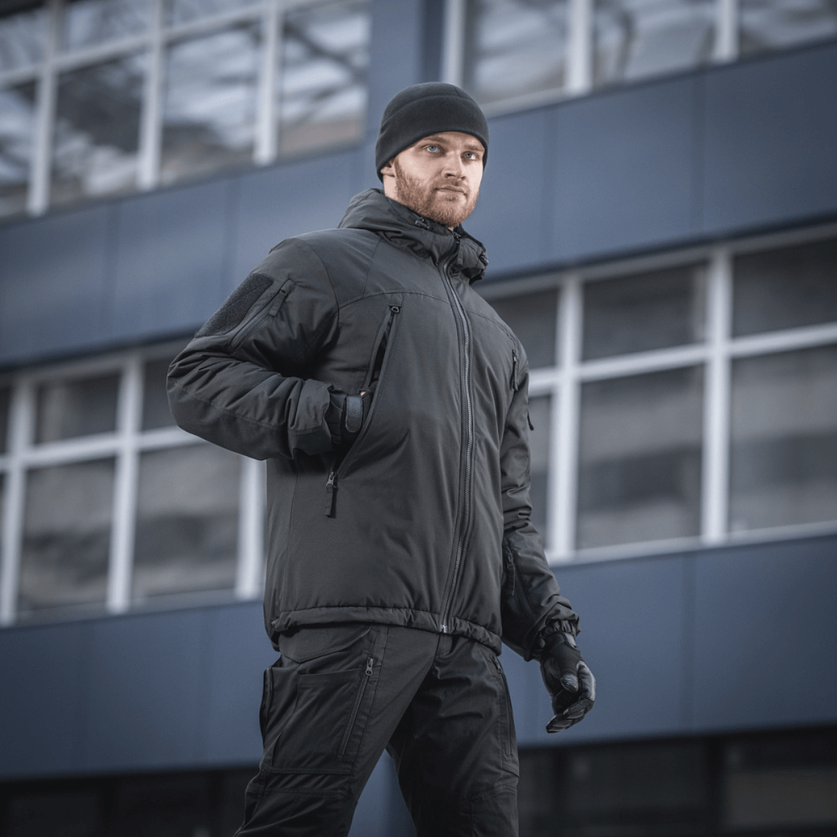 Mand iført M-Tac Alpha Gen.III Pro Vinterjakke i sort, velegnet til temperaturer ned til -20° C, foran moderne bygning.