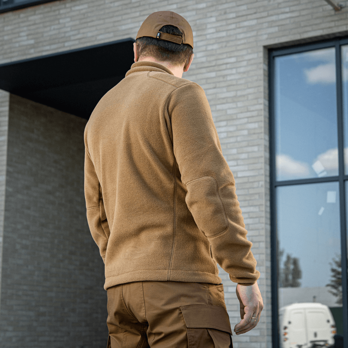 Mand iført M-Tac Nord Polartec Fleecejakke i brun, set bagfra med moderne bygning i baggrunden.