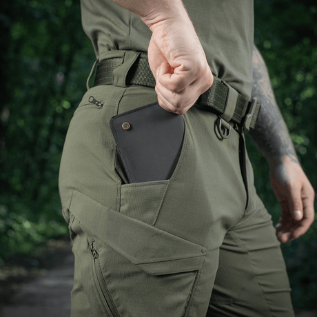 Mand iført M-Tac Aggressor Summer Flex Shorts i skoven med en hånd i lommen, viser mobiltelefonopbevaring. Produktsøgeord: M-Tac Aggressor Summer Flex Shorts.