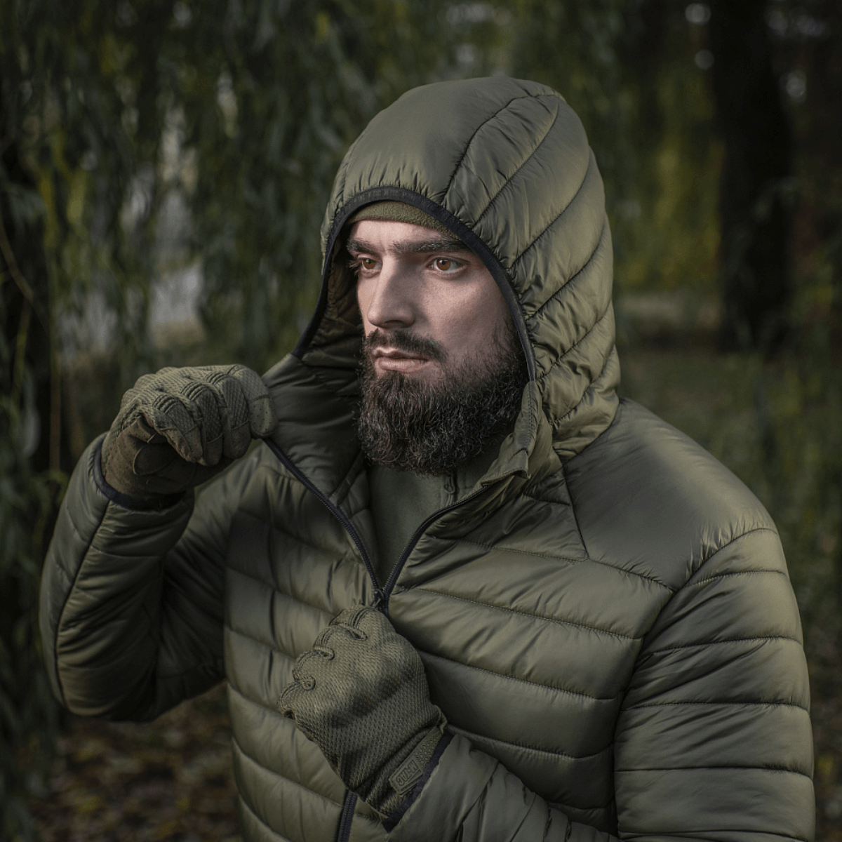 Mand iført M-Tac Stalker Gen.III Jakke i skovmiljø, viser jakke med hætte og lynlås, ideel til overgangssæsonen