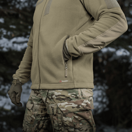 Person iført M-Tac Combat Fleece Polartec Jakke i skovmiljø, med camouflagebukser og handsker, fremhævet varme- og slidstærke egenskaber.