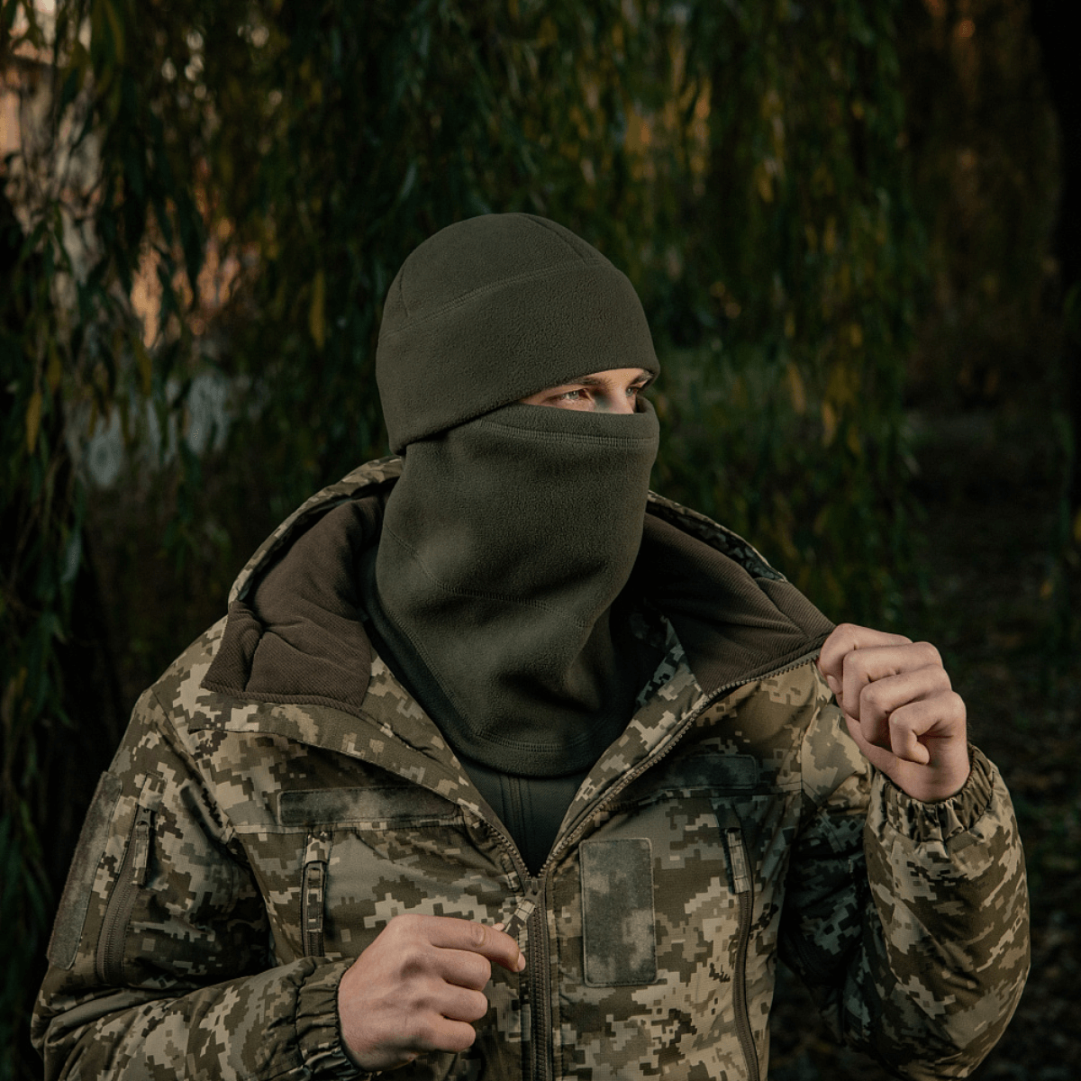 M-Tac Anatomisk Snood Justerbar Elite Fleece (320 g/m²) bæres som balaclava i koldt vejr, set mod baggrund af skov i camouflagejakke.