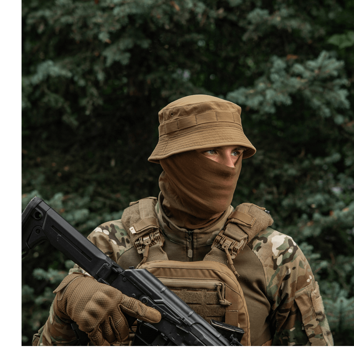 Person i camouflage med hat og maske i skov, bevæbnet med gevær.