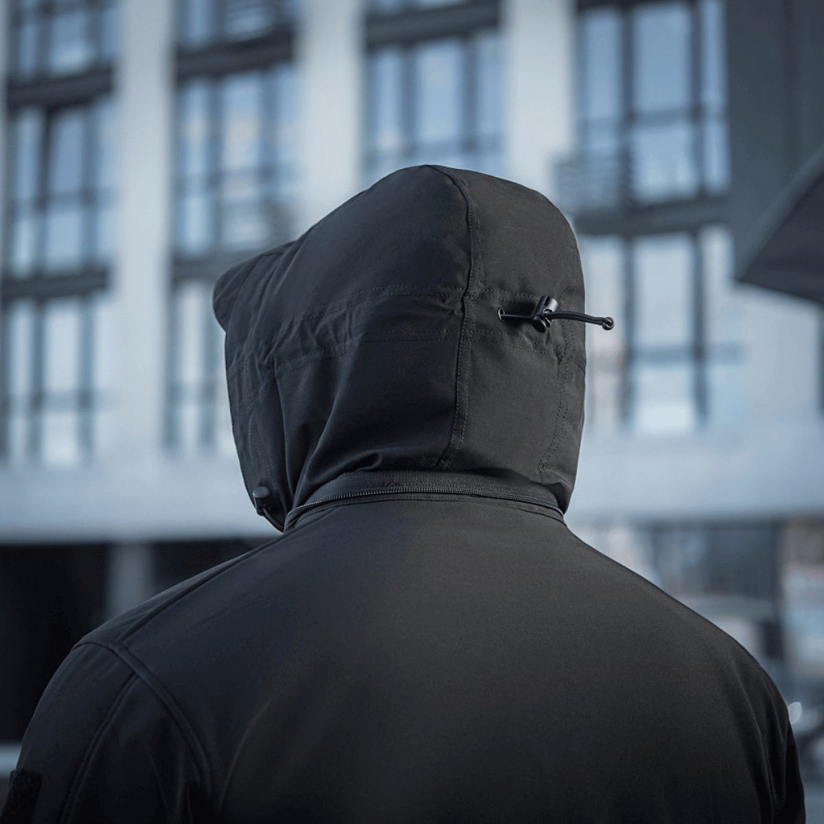 Mand i M-Tac Soft Shell Jakke set bagfra, viser hætte og moderne bymiljø baggrund, ideel til beskyttelse mod dårligt vejr.