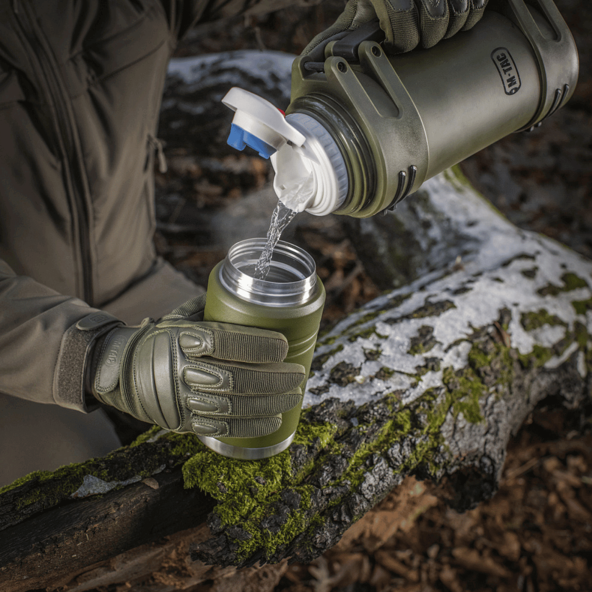 Person hælder vand fra en M-Tac Termoflaske 750 ml med Folde-ske, perfekt til camping og udendørs aktiviteter, på en skovsti.
