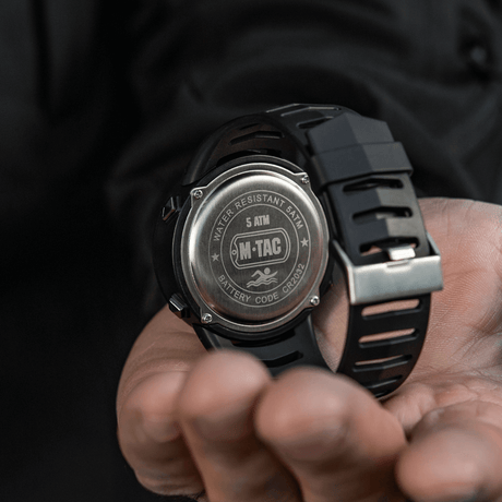 M-Tac Tactical Ur 2147 med stållåg, "WATER RESISTANT 5ATM" og "BATTERY CODE CR2032" vises i en hånd.
