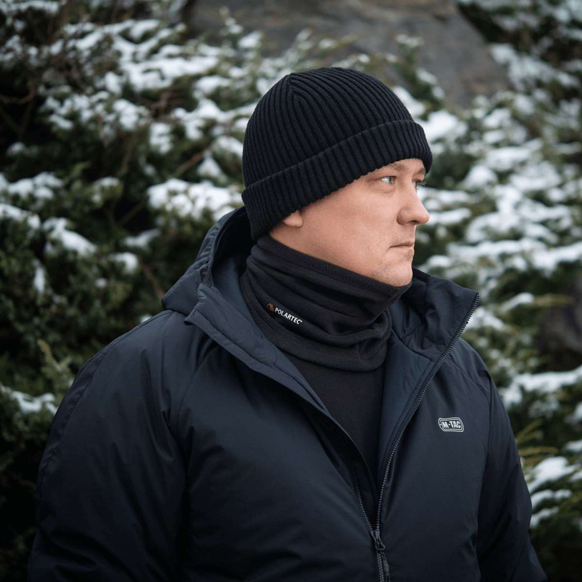 Mand iført M-Tac Tube Scarf Polartec foran sneklædte grantræer; varm multifunktionel halstørklæde til udendørs brug i kolde årstider.