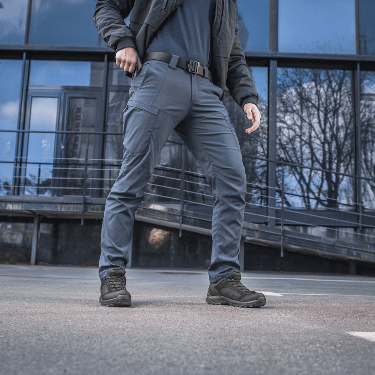 Mand iført M-Tac Bukser Sahara Flex Light, som tilbyder komfort og stil på solrige sommerdage, stående foran en moderne bygning.