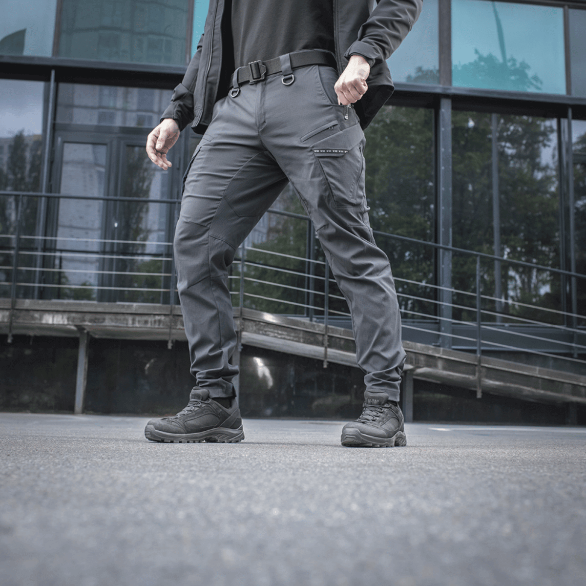 Mand iført M-Tac Bukser Aggressor Summer Flex i Rip-Stop nylon, ideel til varmt vejr, foran moderne bybygning.