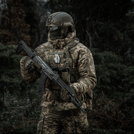 Soldat i camouflage med riffel i skov, bærer M-Tac Patch M67 Granat 3D PVC på taktisk vest.