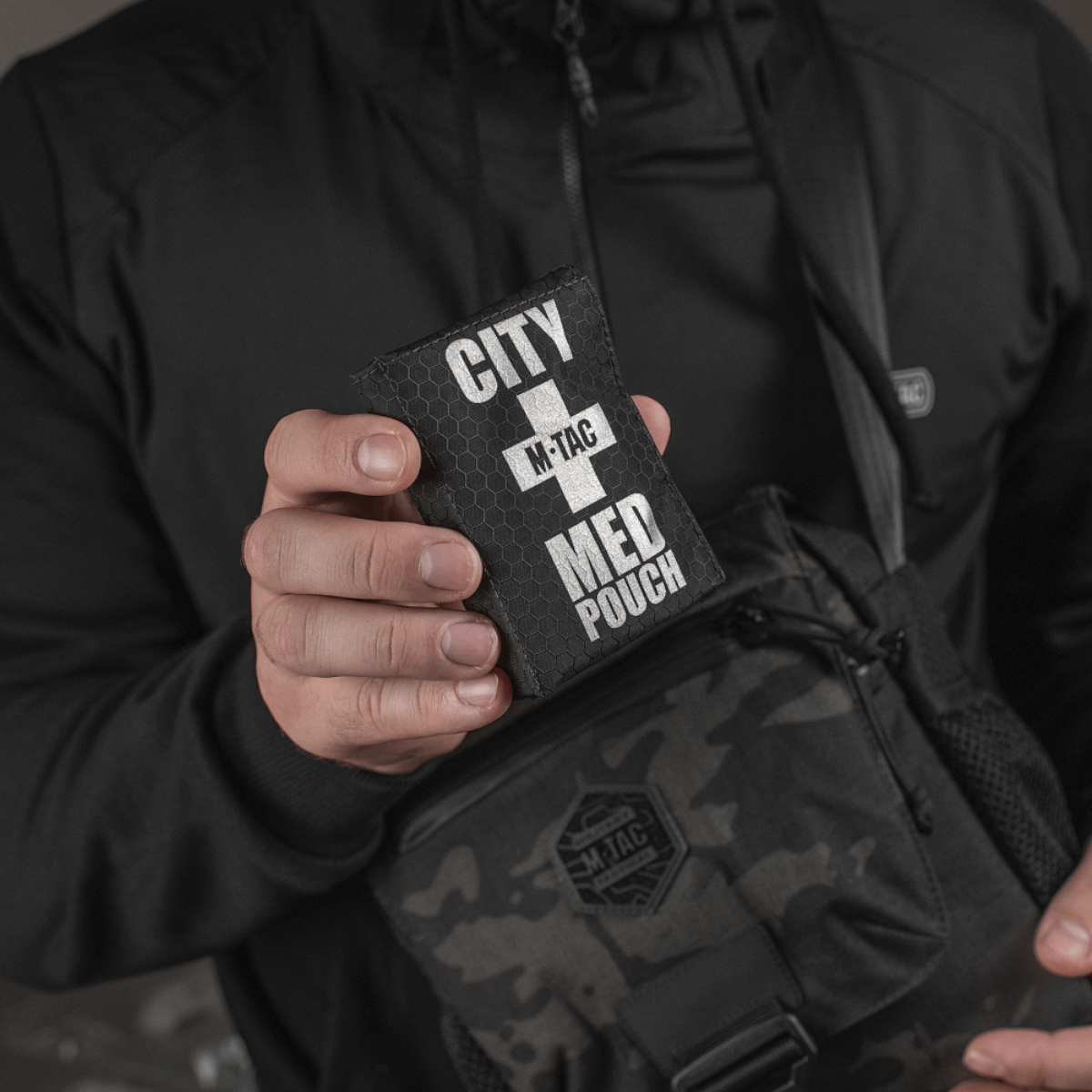 Person holder M-Tac City Med Pouch Hex, et kompakt førstehjælpskit i slidstærk Hex Polyester med M-Tac-logo og velcrolukning.