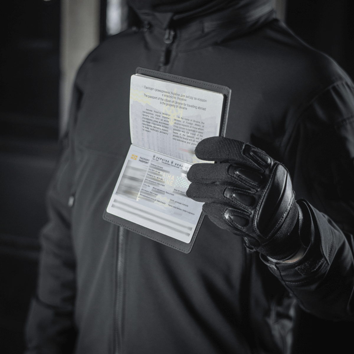 Person holding pasbeskyttelse M-Tac Pascover til ukrainske pas, visende "Passport" tryk, polymerkomposit, sort baggrund.