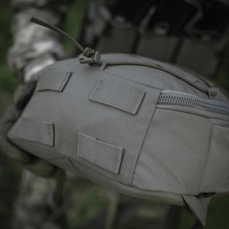 Nærbillede af taktisk rygsæk i robust materiale og beige farve, holdt af en person i camouflageuniform. Fokus på lynlås og stropper.