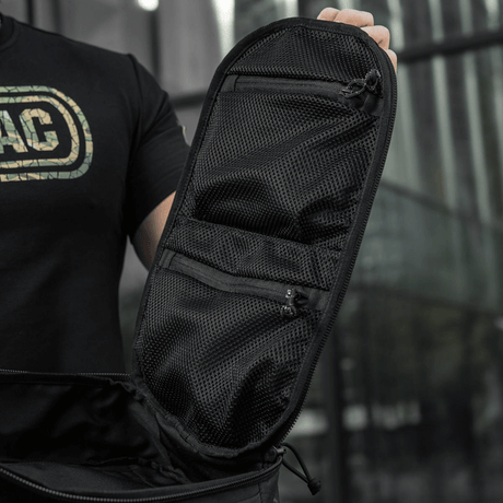 Sort taske med netlommer og lynlåse åbnet på en moderne bybaggrund, betonet af en person i sort trøje.