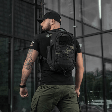 Mand med sort rygsæk og tændstikkamouflage design står foran et moderne glashus.