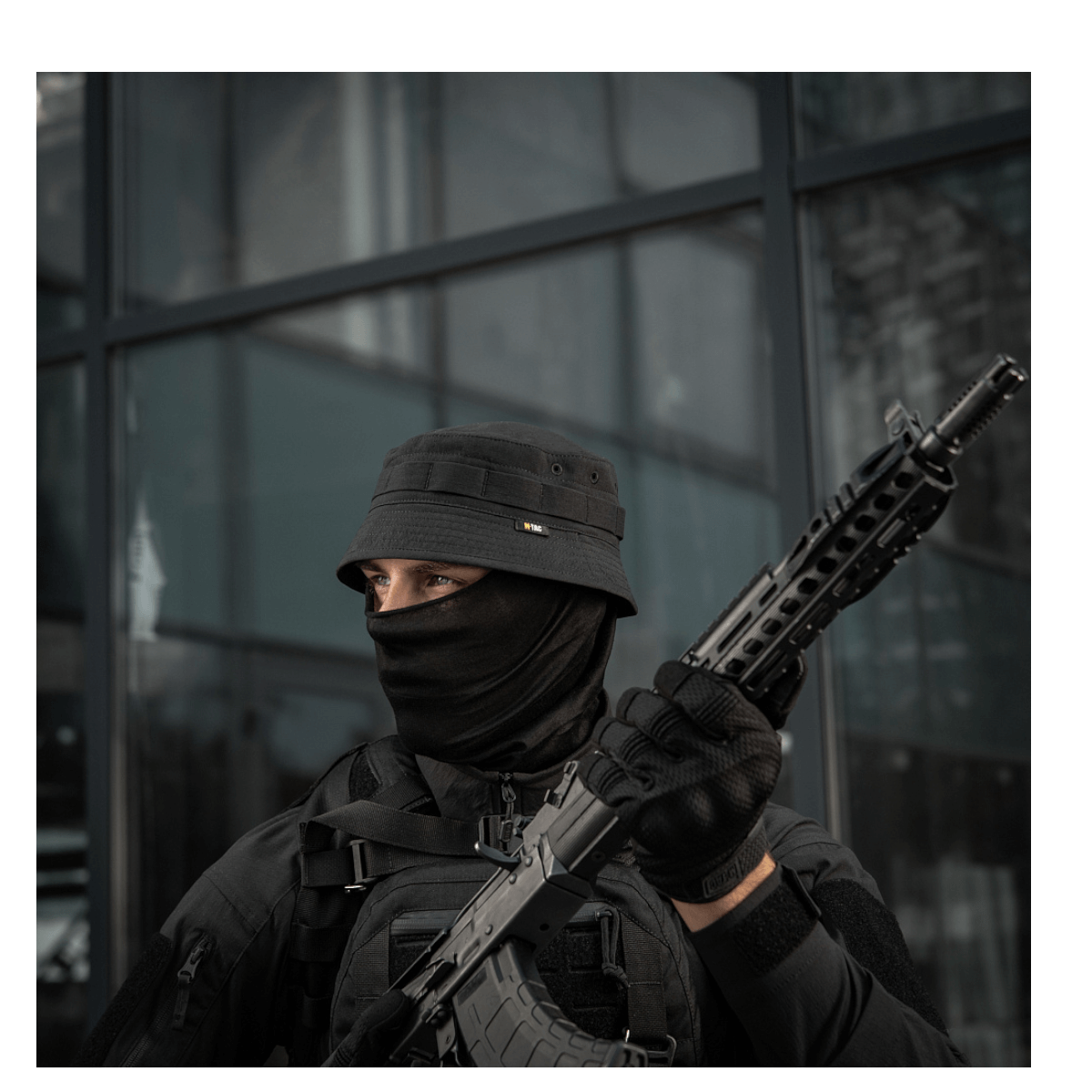 Person wearing tactical gear with a hat and face covering, holding a rifle, standing outside a building.