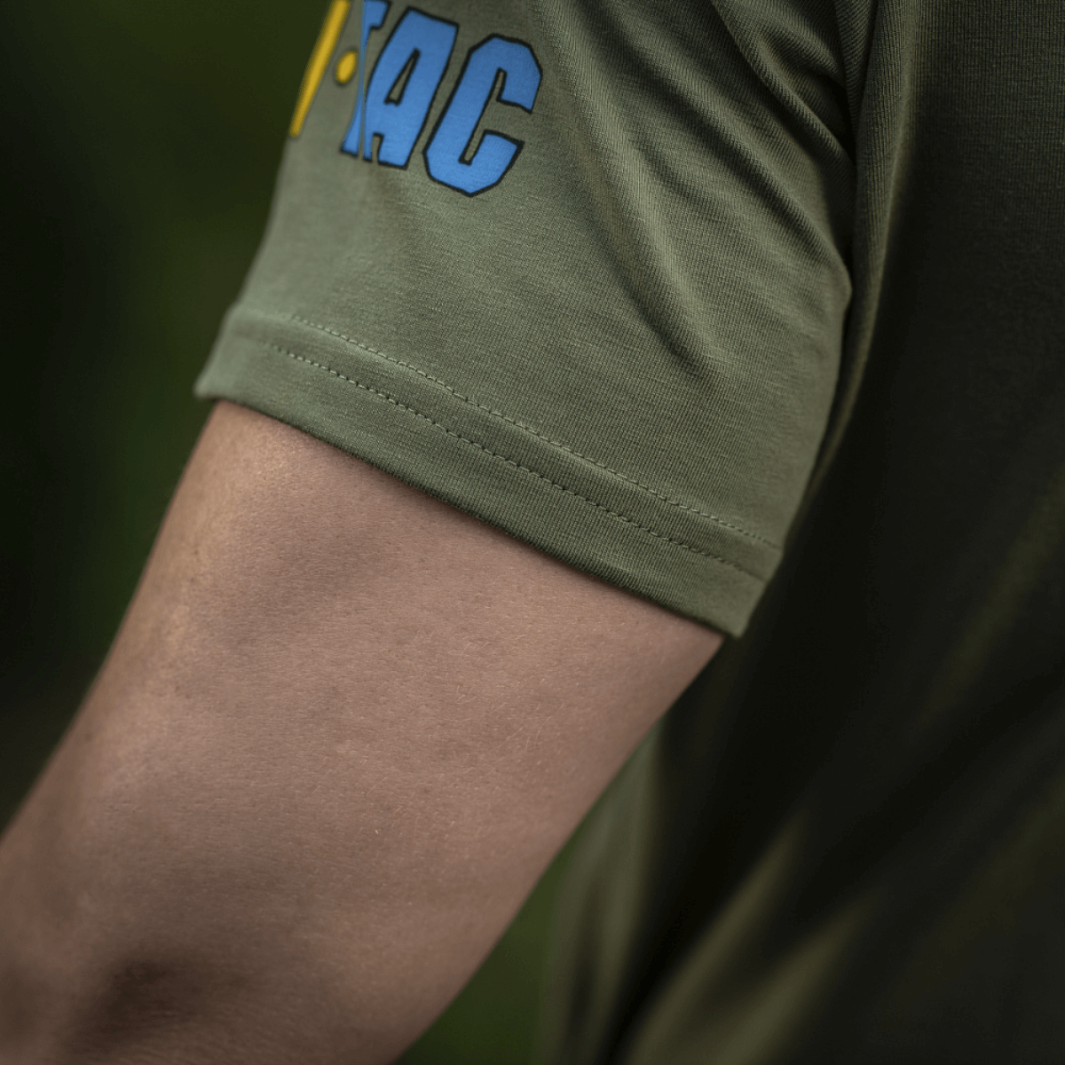 Close-up af ærmet på M-Tac T-Shirt Quadcopter med logo i blå og gul på olivengrøn baggrund, viser bomuld og elastan blanding.