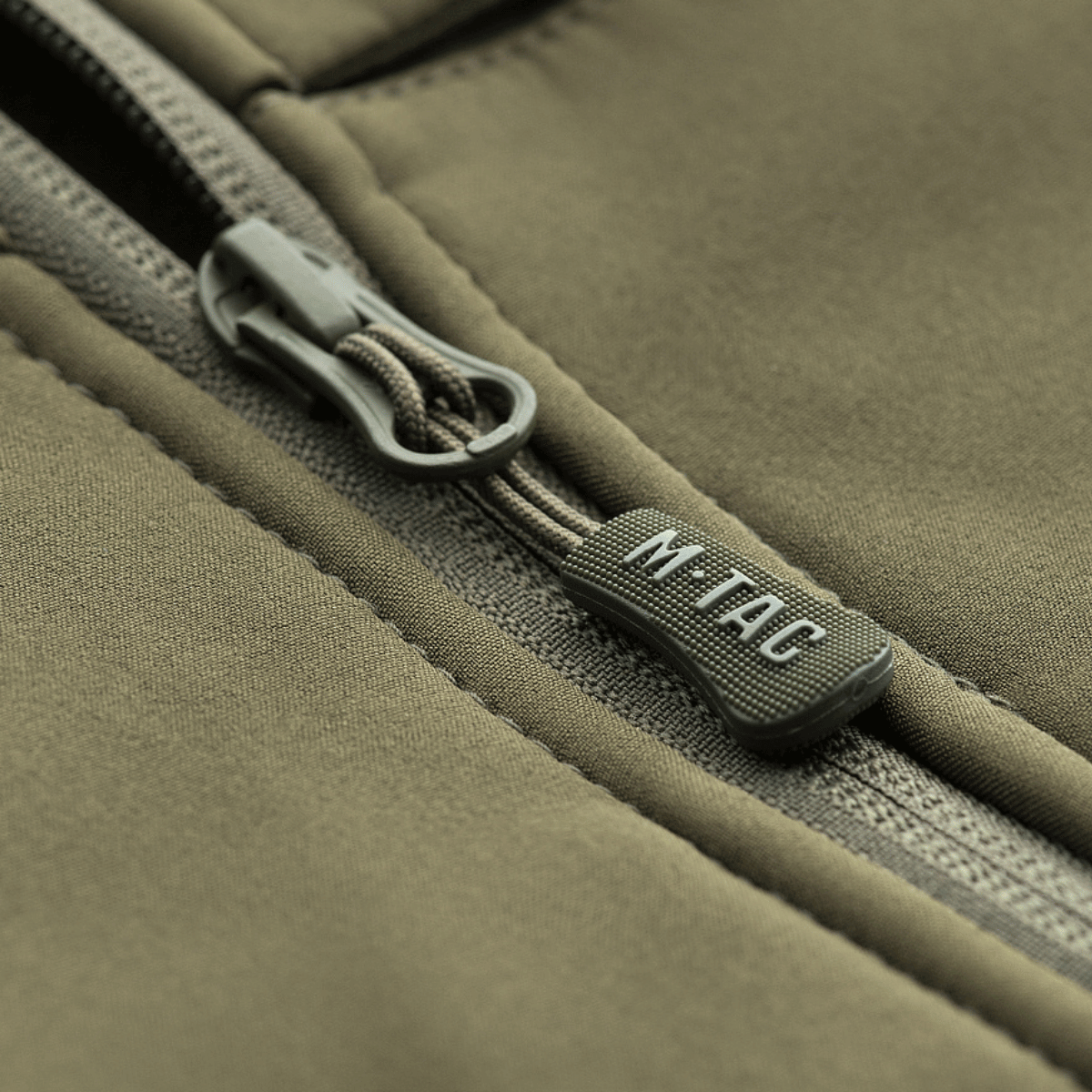 Nærbillede af lynlås på M-Tac Soft Shell Police Jakke med M-Tac logo, designet til beskyttelse mod vind og vejr.
