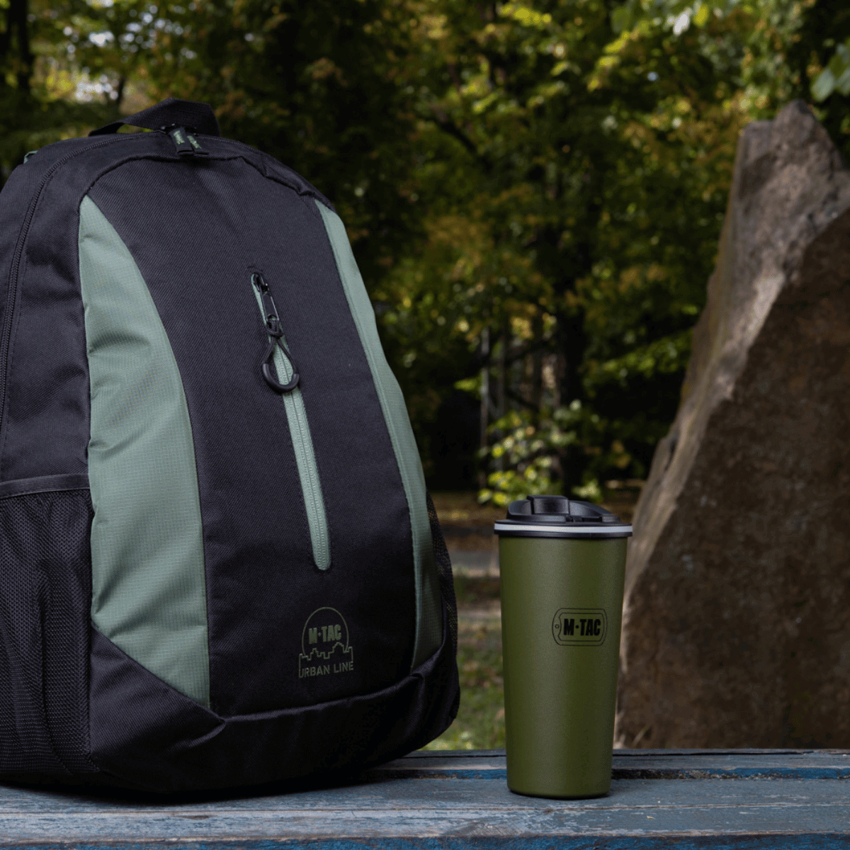 M-Tac Termokop 450 ml med Ventil står ved siden af en rygsæk på et bord i en skovscene.