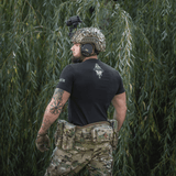 Mand iført M-Tac T-Shirt Odin Mystery foran træer, bagfra, med taktikudstyr. Fokus på figur og komfort i mytologi-inspireret design.