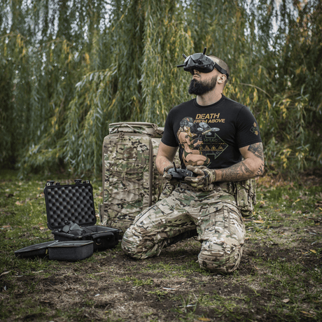 Mand iført M-Tac T-Shirt Delivery Service, inspireret af amerikansk Pin-Up-kultur, styret drone i camouflagetøj i naturen.
