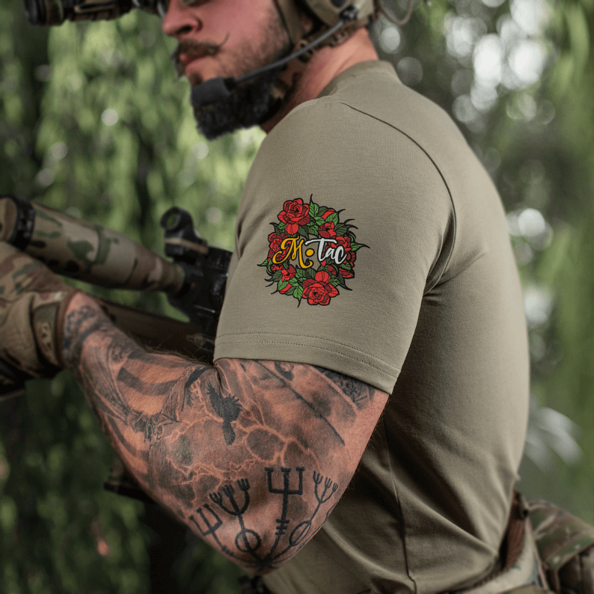 Mand med tatovet arm iført M-Tac T-Shirt Motanka, holder en riffle i naturlig omgivelser, fremhæver slavic inspireret design.